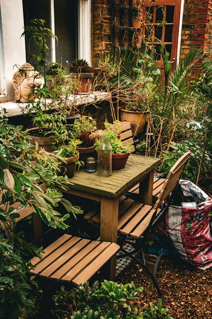 A cozy garden corner adjacent to a red brick house featuring a weathered wooden table surrounded by matching wooden and metal chairs. The table is topped with various potted plants, including succulen