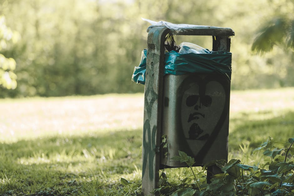 An outdoor scene featuring a metal waste bin with a weathered appearance, situated on a grassy area within a garden. The bin has graffiti art depicting a stylized face with dark sunglasses, a neutral 